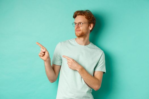 Skeptical caucasian guy with red hair and beard, wearing glasses and t-shirt, looking and pointing left disappointed, judging something bad, standing over mint background.