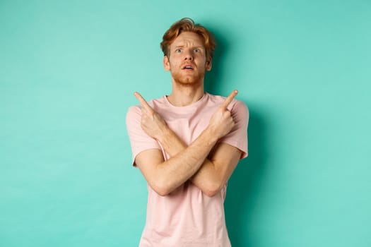 Confused young man in t-shirt looking up, pointing fingers sideways and feeling puzzled, cant decide, standing over turquoise background.