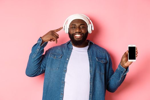 Happy black man listening music, pointing finger at headphones and smiling, showing mobile phone screen app or playlist, standing over pink background.