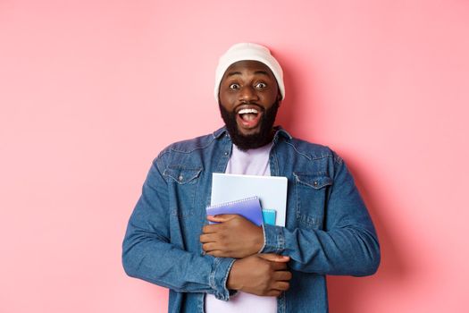 Image of adult african-american man holding notebooks and smiling, studying at courses, standing over pink background.