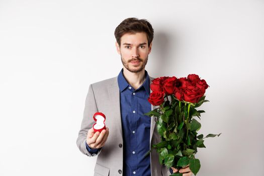 Handsome man in suit going to make a proposal, standing with red roses flowers and engagement rin in box, making romantic surprise, white background.