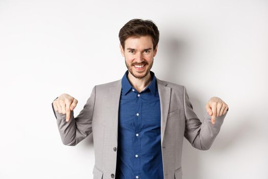 Smiling handsome man in business suit pointing fingers down, showing good deal, look here gesture, standing on white background.