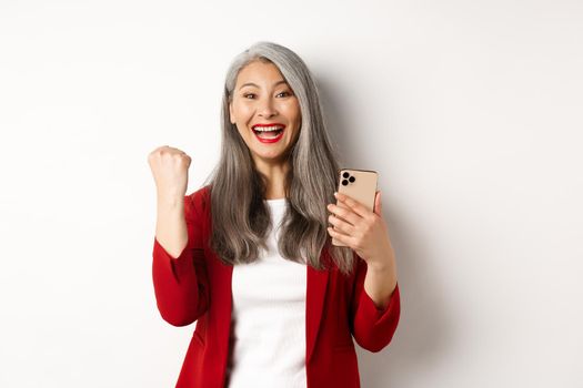 Asian old woman winning online, holding smartphone and making fist pump gesture to celebrate win, triumphing and smiling, standing over white background.