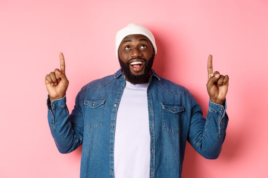 Happy african american man showing advertisement, smiling amazed and staring at camera, pointing fingers up, pink background.