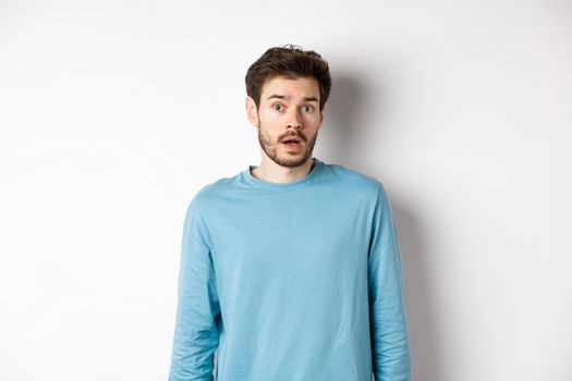 Confused caucasian guy drop jaw and stare startled at camera, standing over white background.