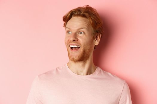 Close-up of handsome caucasian guy with red hair and beard, looking left with happy smile, checking out promo offer, standing over pink background.
