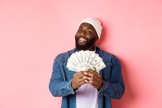 Satisfied rich african-american man showing money, smiling pleased and show-off with income, standing with dollars over pink background.
