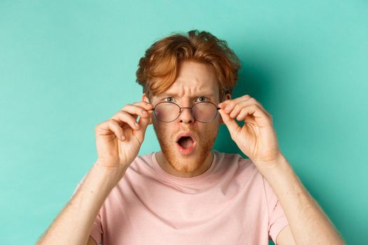 Close up portrait of redhead guy take-off glasses and gasping startled, looking shocked at camera, standing over turquoise background.