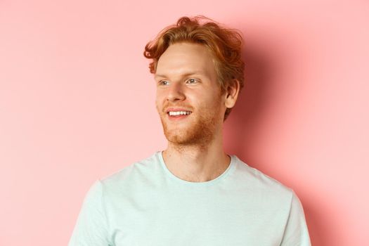 Portrait of attractive caucasian guy with red messy hair and beard, turn head and looking left with pleased smile, standing over pink background.