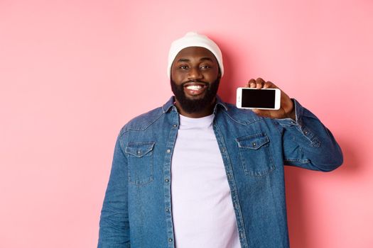 Online shopping and technology concept. Handsome Black man showing smartphone screen horizontally and smiling satisfied, recommending app, pink background.