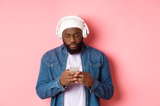 Man looking serious while reading messagin on phone, listening music in headphones, standing over pink background.