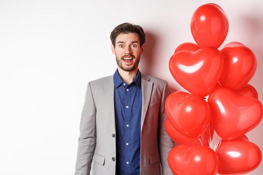 Lovers day. Excited handsome man in suit standing near red hearts balloons, raising eyebrows and looking surprised, standing over white background.