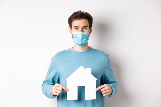 Covid and real estate concept. Man searching for flat, showing paper house cutout, wearing medical mask, standing on white background.