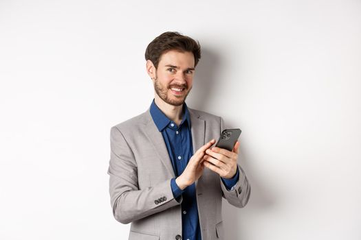 Smiling successful business man using smartphone on white background, standing in suit.