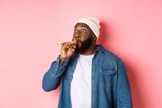 Funny Black man blowing in party whistle and pouting, staring sideways with popped eyes, standing against pink background.