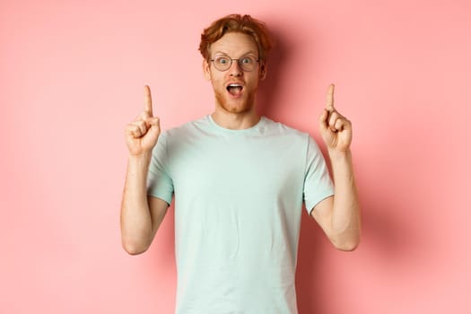 Portrait of surprised guy checking out advertisement, gasping amazed and pointing fingers up, showing promo, standing over pink background.