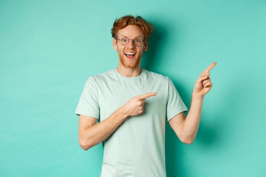 Handsome smiling man with red hair and beard, looking amused and pointing at upper right corner, showing promo offer, standing over mint background.