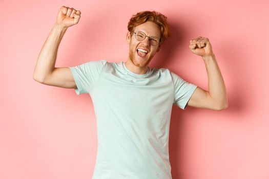 Cheerful young man with red hair looking happy, raising hands up in fist pumps gesture, celebrating success, feel like champion, winning and standing over pink background.