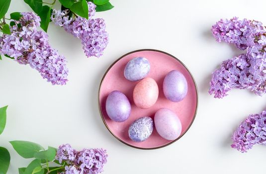 Close-up top view of beautiful nacreous pink and lilac easter eggs lie on a pink plate framed by flowering branches of lilac on a white background. Selective focus.