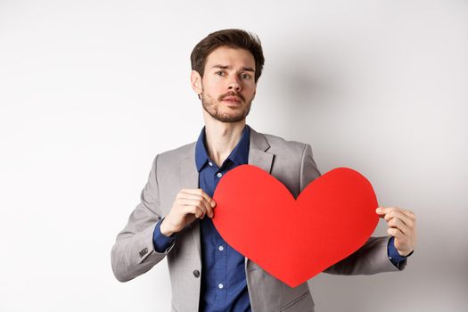 Passionate handsome man showing heart pounding gesture with red valentines cutout, standing in suit and searching for love, standing over white background.