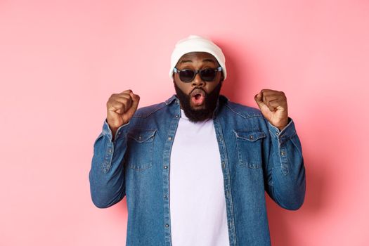 Excited african-american guy in sunglasses and beanie winning and rejoicing, making fist pump in triumph, staring at camera and celebrating, standing over pink background.