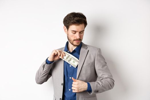 Successful businessman put dollars inside suit pocket, hiding money, standing on white background.