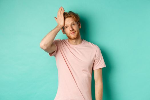 Annoyed and bothered redhead male model showing facepalm gesture, standing over mint background.