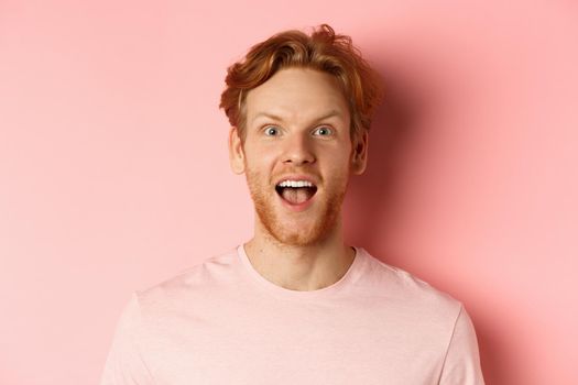 Headshot of amazed redhead male model in t-shirt open mouth, looking fascinated and happy at camera, standing over pink background.