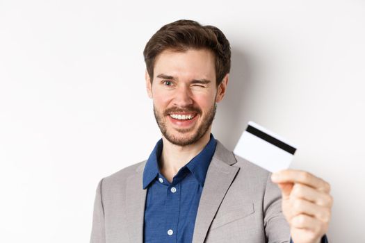 Shopping. Cheerful bearded guy in suit winking and smiling, showing plastic credit card at camera, white background.