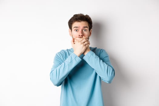 Image of shocked young man staring startled and covering mouth with hands, hear gossips, standing on white background.