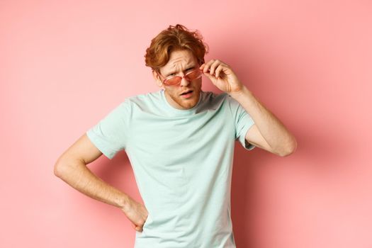 Tourism and vacation concept. Confused funny guy with red messy hair and beard, take-off glasses and frowning puzzled, standing over pink background.