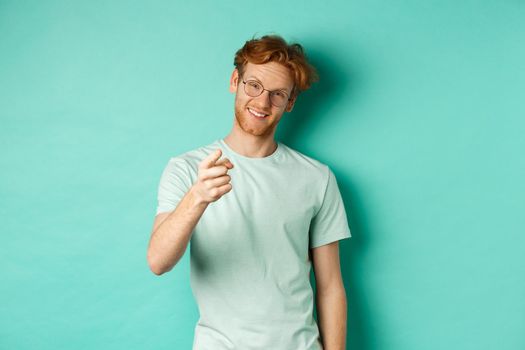 Handsome young man with ginger hair, wearing glasses and t-shirt, pointing finger at camera and smiling, choosing you, making invitation, standing over turquoise background.