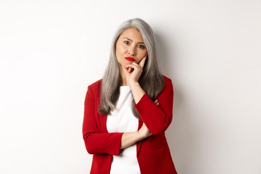 Annoyed and bothered asian businesswoman in red blazer, pouting and looking irritated at camera, standing over white background.