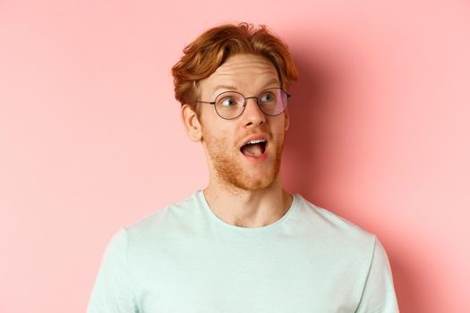 Headshot portrait of amazed redhead man in glasses, open mouth and gasping, looking at upper right corner banner or logo, standing over pink background.