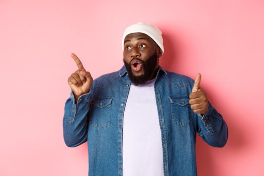 Happy african-american man showing thumb-up and pointing at upper left corner copy space, praising good deal, standing over pink background.