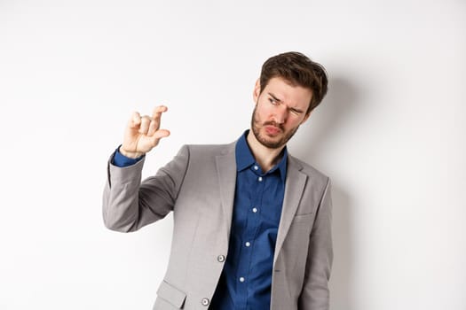 Businessman trying to see something tiny, showing small thing size with fingers, standing in suit on white background.