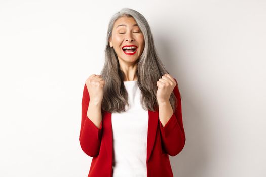 Relieved asian senior businesswoman making fist pump, saying yes and smiling satisfied, triumphing and winning, standing over white background.