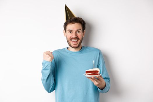 Celebration and holidays concept. Cheerful young man celebrating birthday in party hat, saying yes and fist pump in joy, holding bday cake, white background.