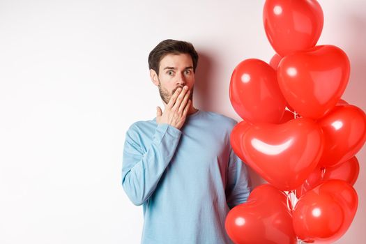 Portrait of man boyfriend standing near Valentines day heart balloons and gasping shocked, standing over white background concerned.
