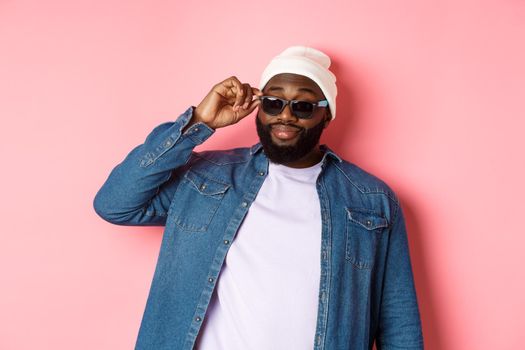 Cool and sassy african-american bearded man, looking confident, touching sunglasses and stare at camera please, standing over pink background.
