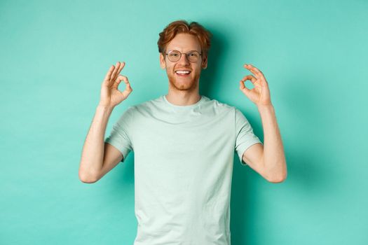 Attractive male model with red hair, wearing glasses, showing OK sign in approval and saying yes, smiling satisfied, standing over mint background.