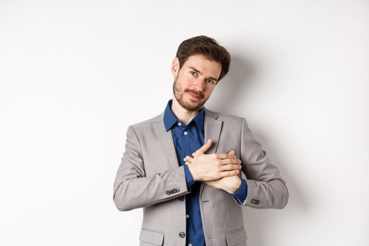 Romantic handsome guy with beard holding hands on heart and smiling, thanking you, feeling nostalgic, having tender memories in heart, white background.