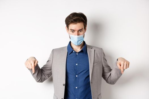 Covid-19, pandemic and business concept. Shocked guy in medical mask and office suit pointing, looking down startled, white background.