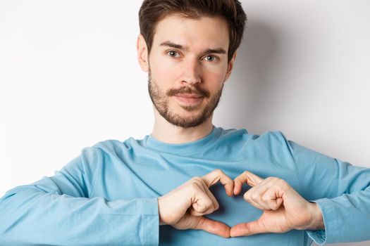 Close-up of young romantic man with beard, showing heart gesture and looking at camera, show I love you sign, white background.