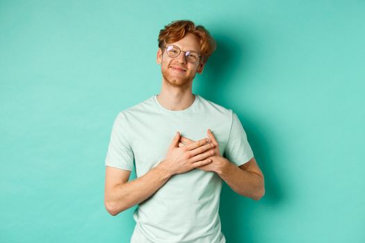 Handsome young man with red hair and glasses, holding hands on heart and smiling, saying thank you, feeling grateful and touched, standing over turquoise background.