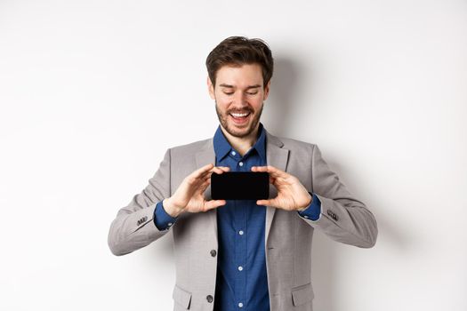 E-commerce and online shopping concept. Happy young man in suit laughing, showing empty smartphone screen horizontally, wite background.