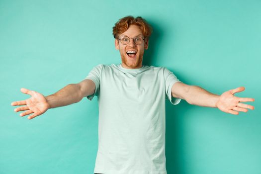 Excited young redhead guy in glasses strethced out hands in warm welcome, invite you and smile friendly at camera, standing happy over turquoise background.