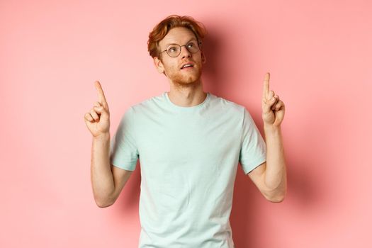 Dreamy young man with red hair and beard pointing fingers up, looking pensive at top, standing against pink background.
