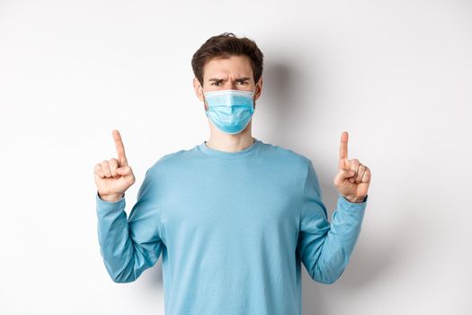 Coronavirus, health and quarantine concept. Disappointed young man frowning at absurd, pointing fingers up and complaining at bad promo, wearing medical mask, white background.