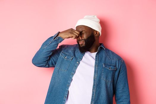 Disgusted african-american man shut nose from aversion, displeased by awful bad smell, standing in beanie and denim shirt over pink background.